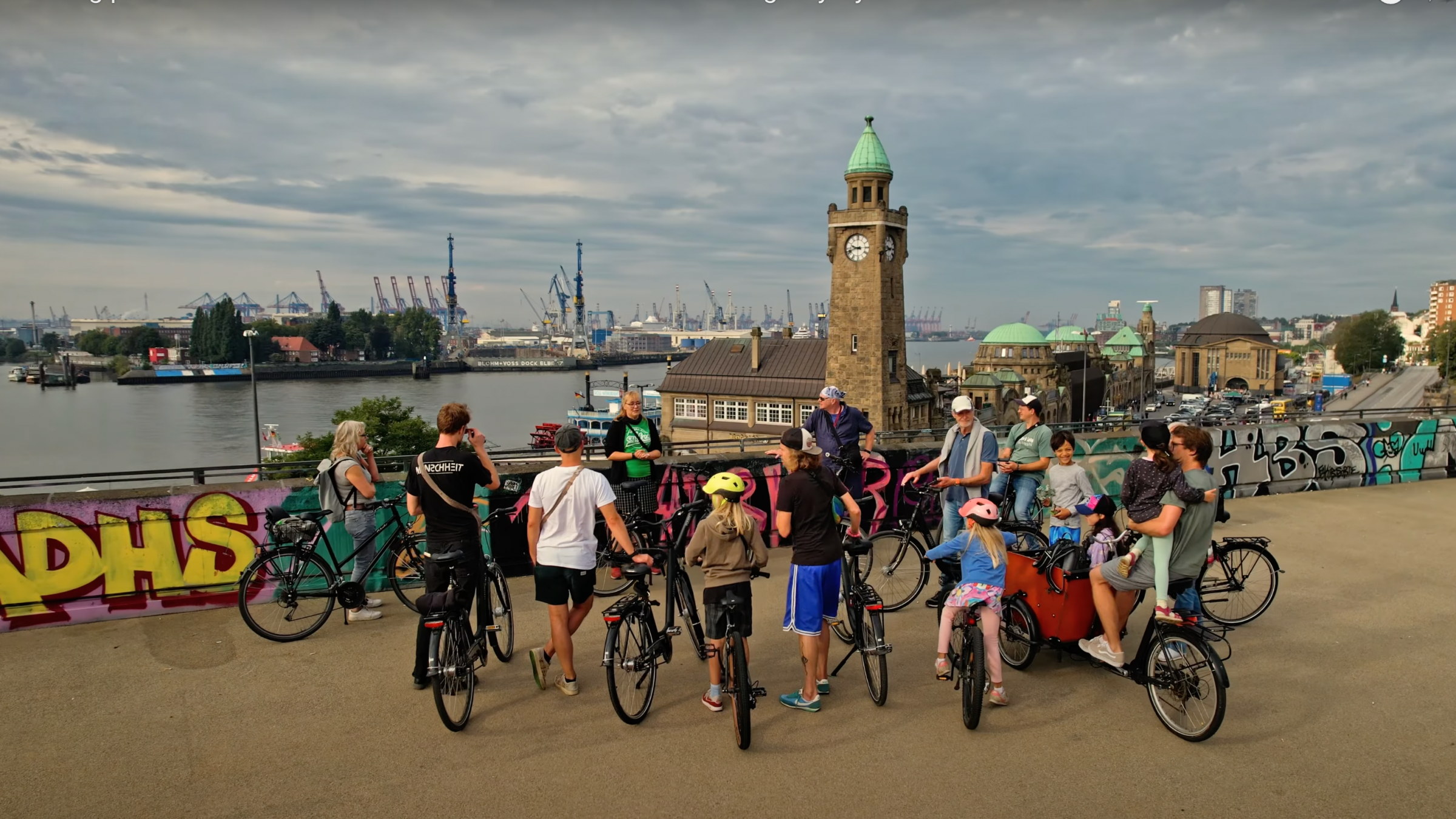 Cyclists gather on a promenade, with a clock tower and industrial harbor in the background.