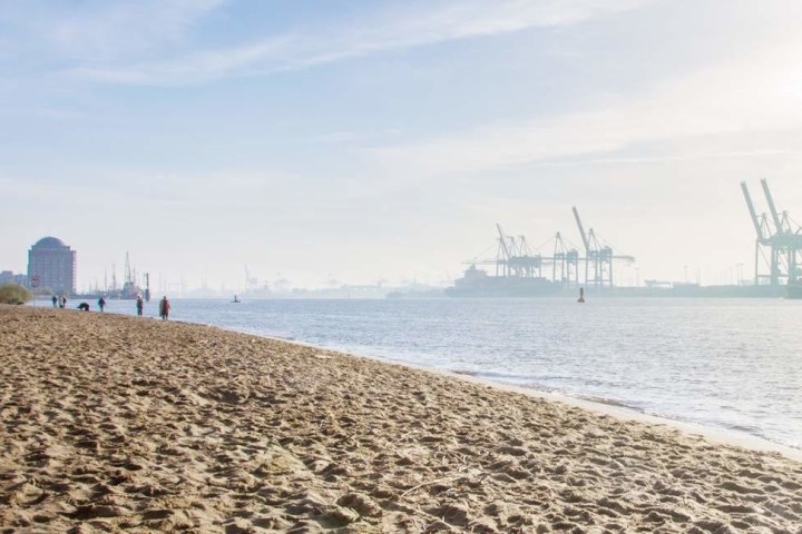 Sandy beach with people walking, industrial port with cranes in background on a sunny day.