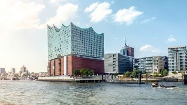 Modern waterfront building with wave-like roof by a cityscape and water.