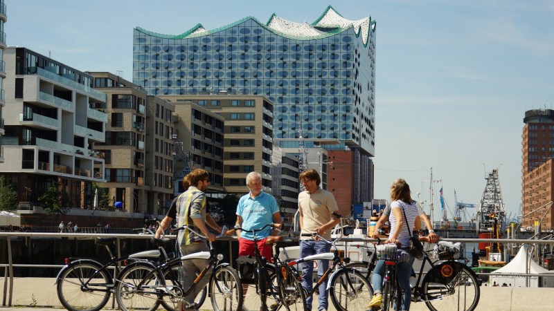 Four people with bicycles standing in front of modern buildings near a waterfront.