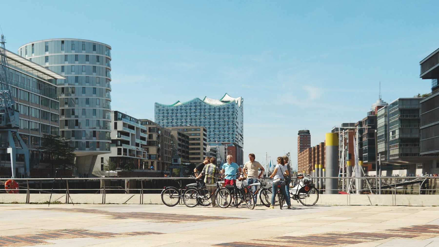 Group of people with bicycles on a plaza by modern buildings under a blue sky.