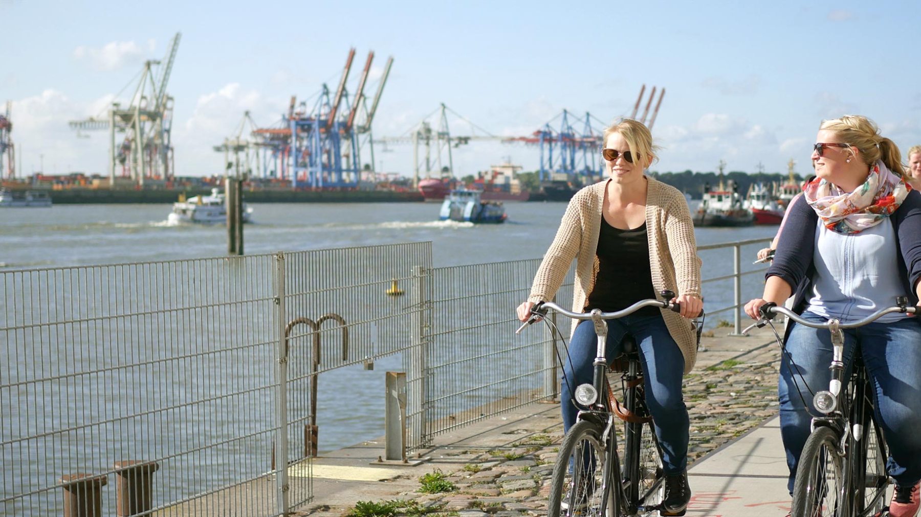 People cycling along a waterfront path with cranes in the background.