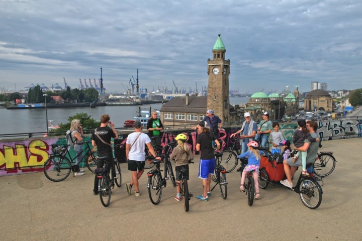 Group of cyclists gathered near graffiti, overlooking river and clock tower on cloudy day.