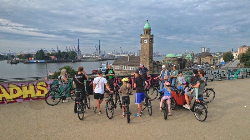 Group of cyclists gathered near graffiti, overlooking river and clock tower on cloudy day.