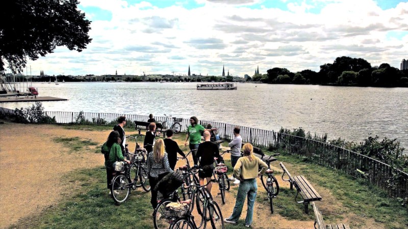 Group of people with bicycles by a lake under a cloudy sky.