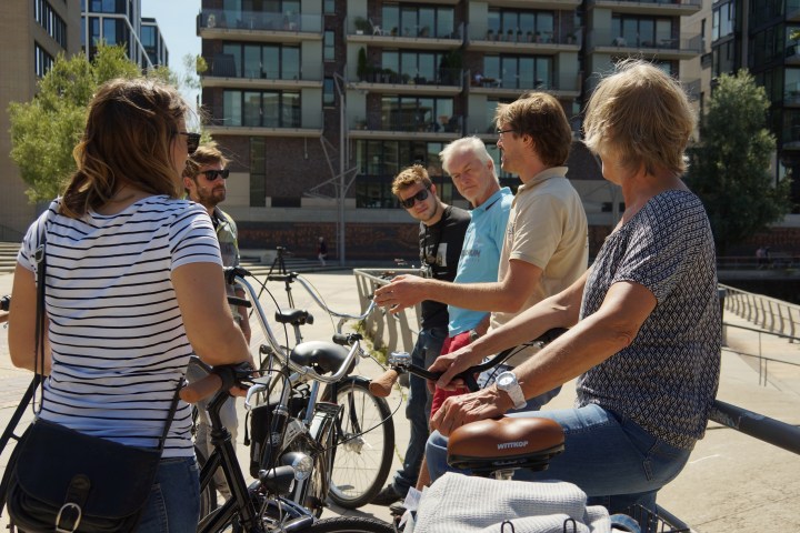 Group of six people with bicycles talking in an urban area with modern buildings.