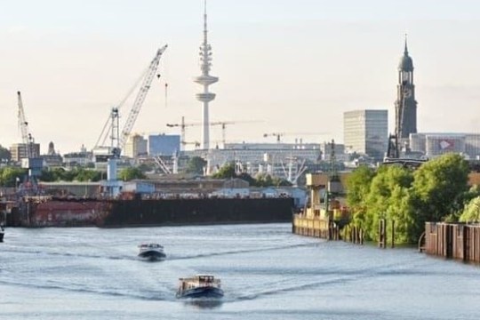 River scene with boats, cranes, green trees, and a distant skyline featuring a tall tower and buildings.