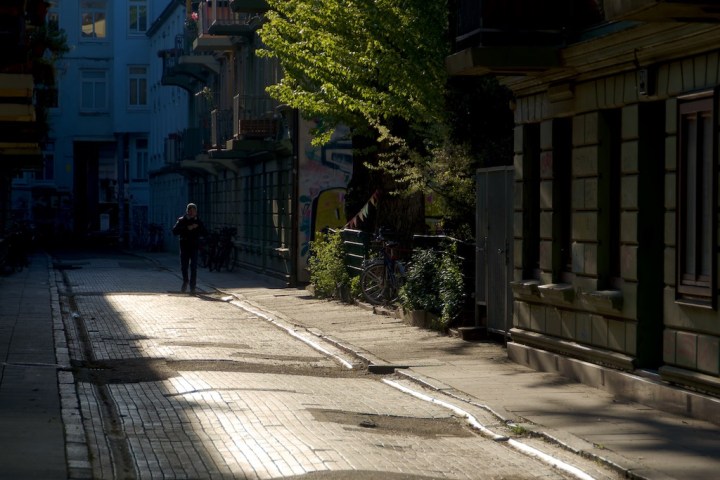 Sunlit alley with person walking, flanked by buildings and trees.
