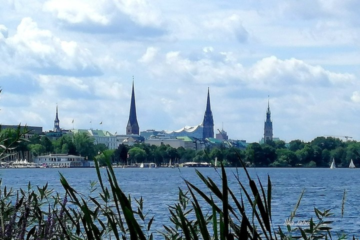 Lakeside view with tall church spires, green trees, and sailboats under a cloudy sky.