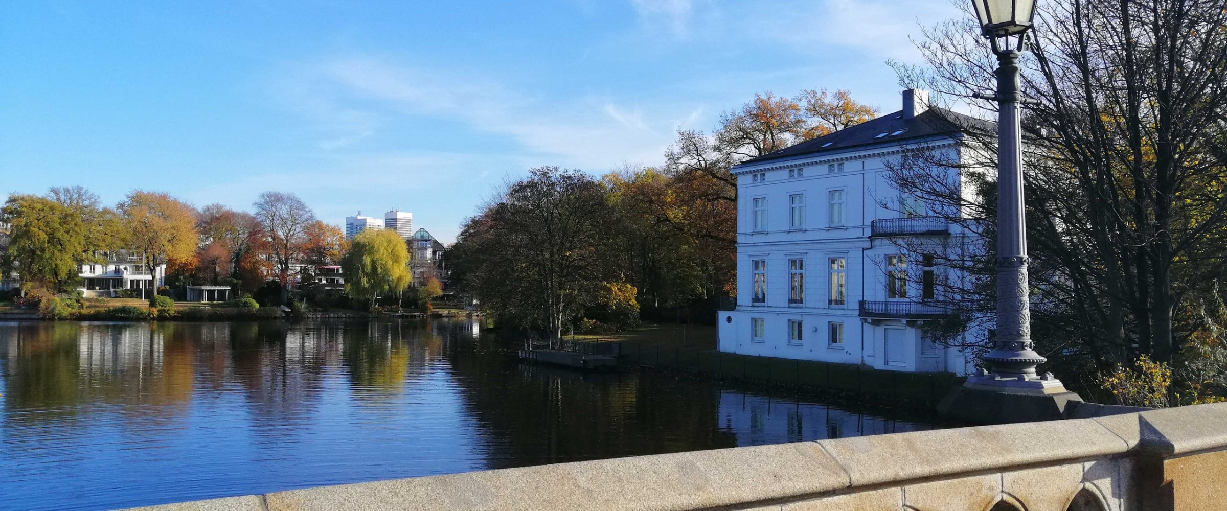 Bridge over water with trees, a white building, and a lamppost under a clear blue sky.