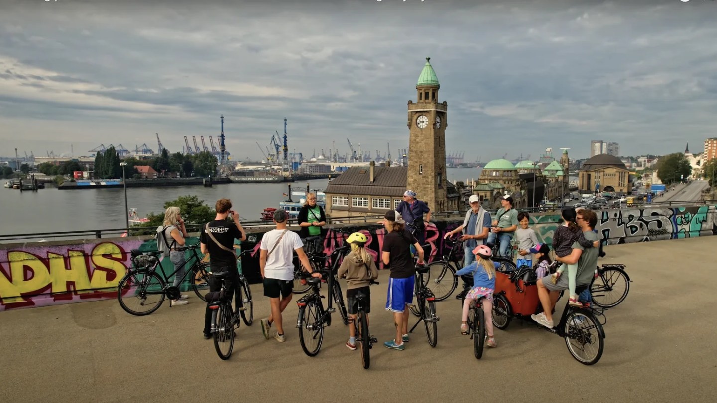 Radtour-Gruppe mit Fahrrädern oberhalb der Hamburger Landungsbrücken, dahinter der Uhrturm und der Hafen.